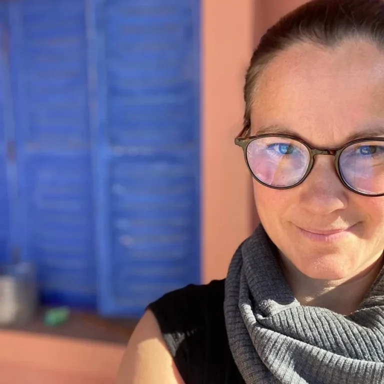 Vanessa Lagnach mit Brille und Schal steht vor einer blauen Fensterläden-Wand.