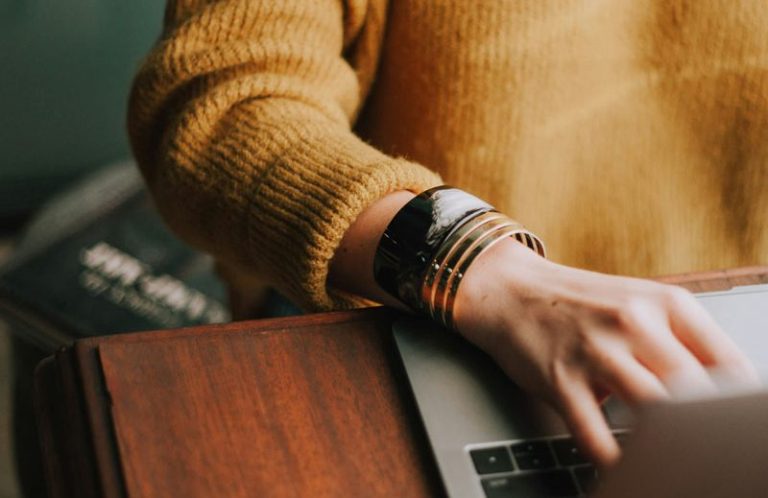 Eine Frau tippt auf einem Laptop, trägt einen gelben Pulli und mehrere Armbänder.