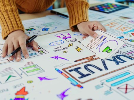 Hände zeichnen bunte Skizzen und Notizen auf einem Tisch voller Papier und Farben.