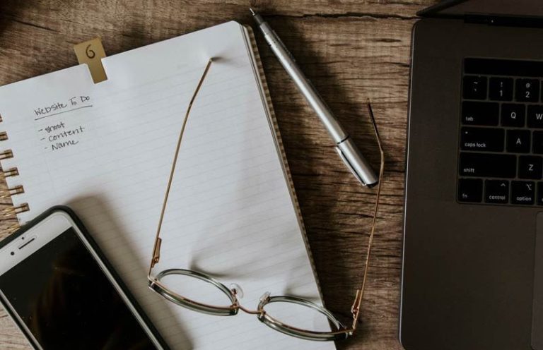 Schreibtisch mit Notizbuch, Smartphone, Brille, Stift und Laptop, um damit eine Website zu planen.